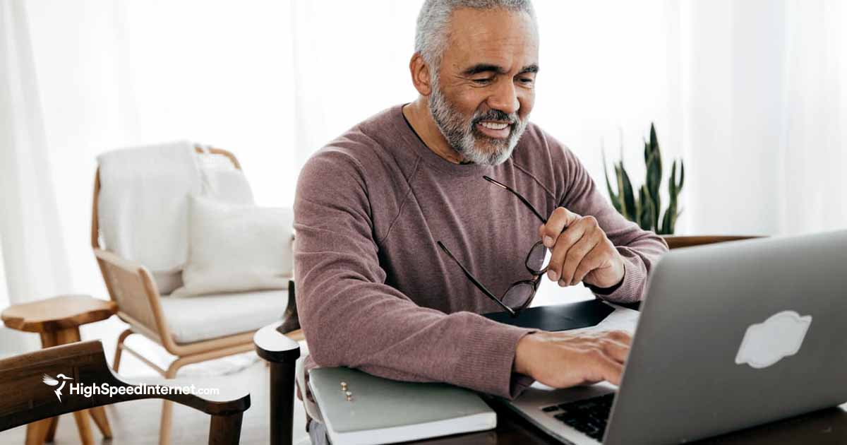 friendly elderly man sitting at a desk using laptop while holding glasses in his hand
