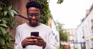 happy black male using cell phone next to wall outdoors with foliage in the afternoon