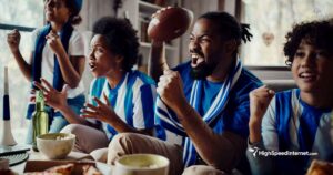 Excited family of four watching a football game eating snacks in a living room with bright windows behind